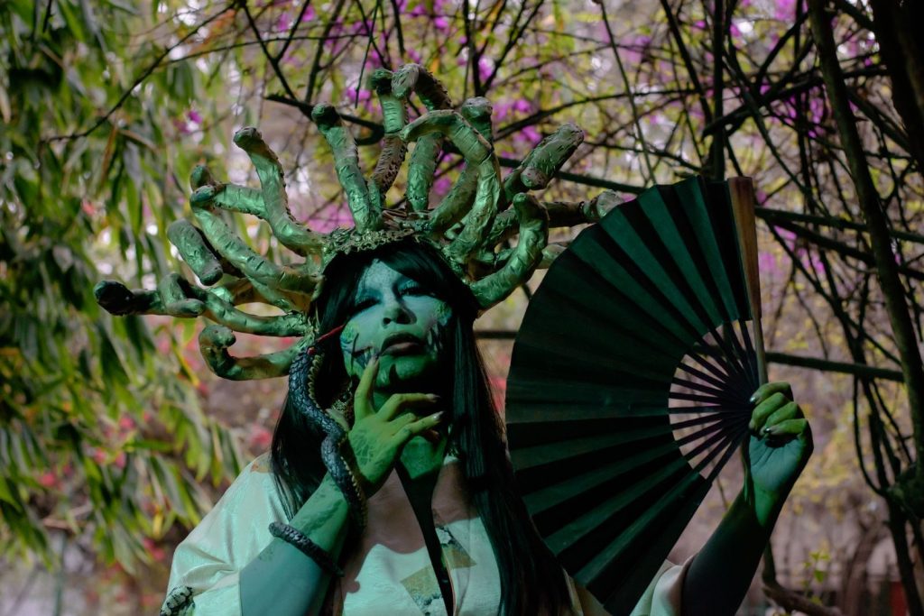 A person in a Medusa costume holding a fan, surrounded by foliage, outdoors in Mexico City.