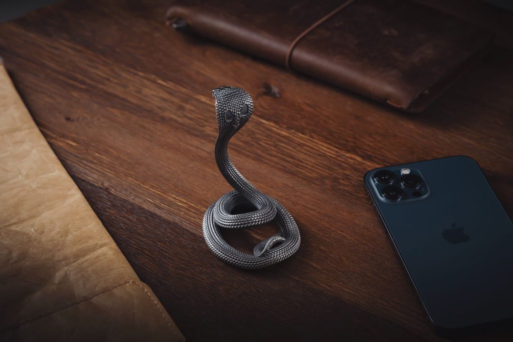 A detailed metallic snake figurine placed on a wooden desk beside a smartphone and a leather notebook.