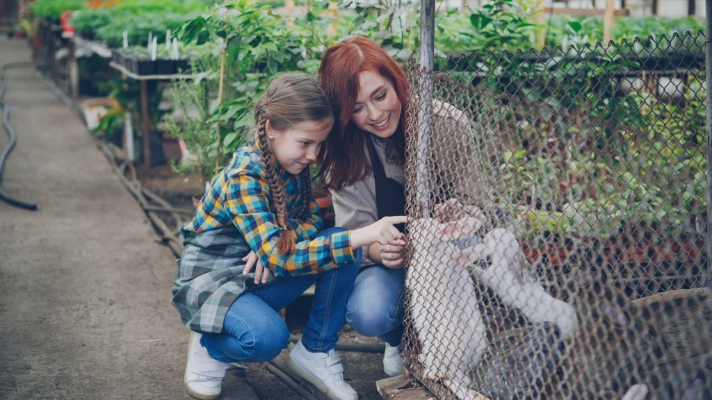 A mother and daughter interact with animals in a sunny greenhouse environment.