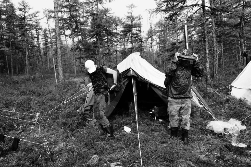 Black and white image of campers setting up a tent in a dense forest, illustrating adventure.