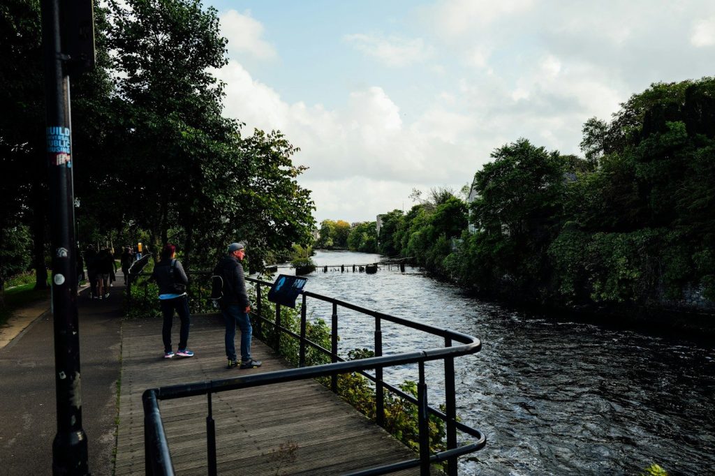 People walking along a river path with trees.