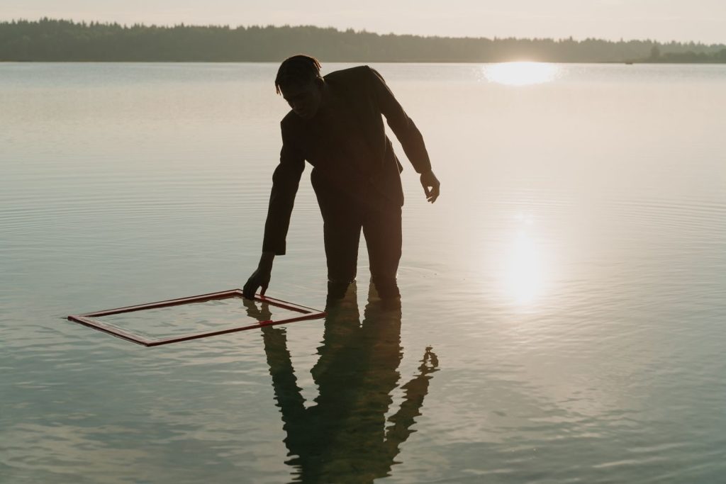 A man in a lake holding a frame at sunset, reflecting on the calm water surface.