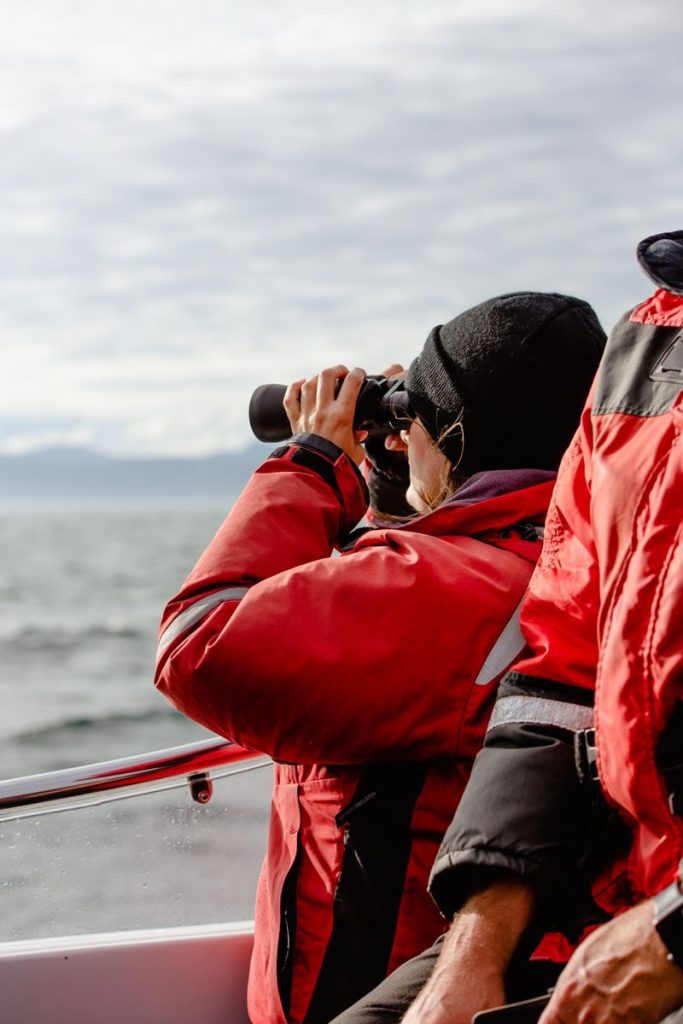 A person in red jacket uses binoculars to observe the ocean from a boat, capturing adventure and exploration.