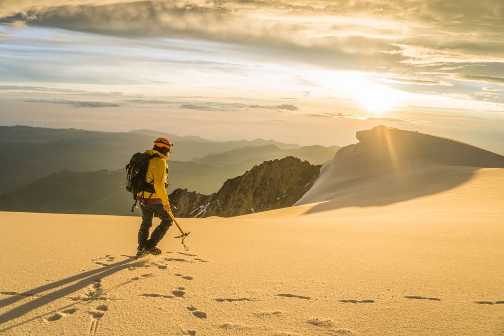 A lone mountaineer walks through a snow-covered mountain range during a golden sunrise.