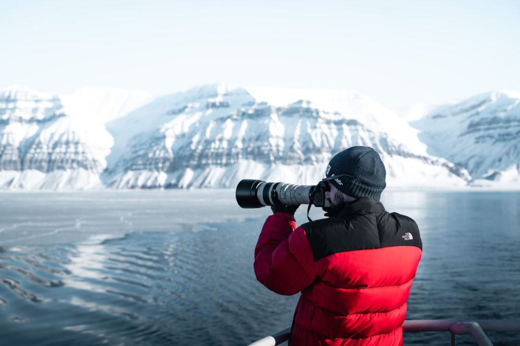 A man in a red jacket photographs snow-covered mountains across a lake. Winter landscape.