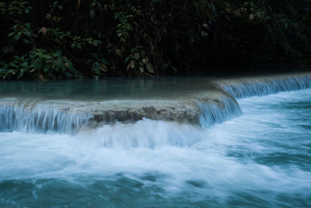 A tranquil waterfall flows over a rocky ledge surrounded by lush green foliage in a serene forest setting.