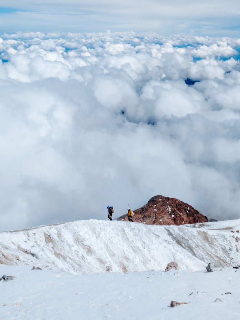 Two hikers exploring a snowy mountain peak surrounded by clouds and sky.