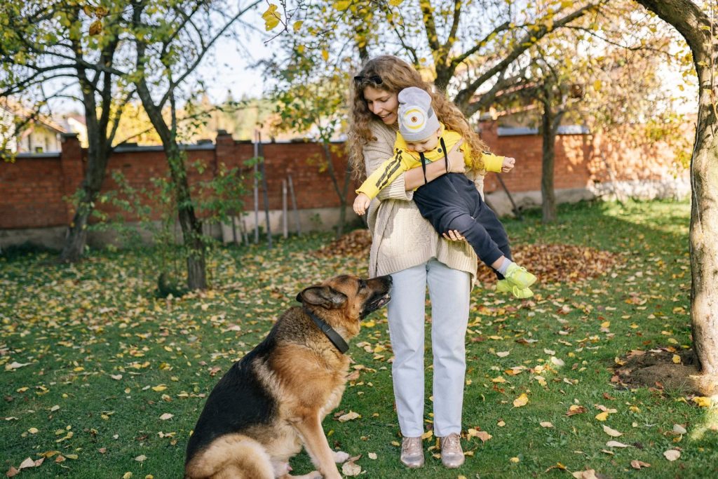 A mother holds her child in a cozy yard while a dog sits nearby amidst the fall foliage.