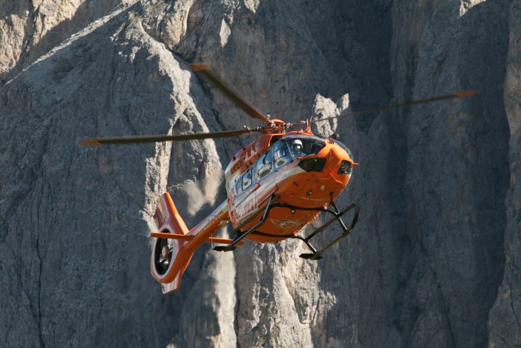 A bright orange rescue helicopter flying amidst the rugged Dolomites mountains.